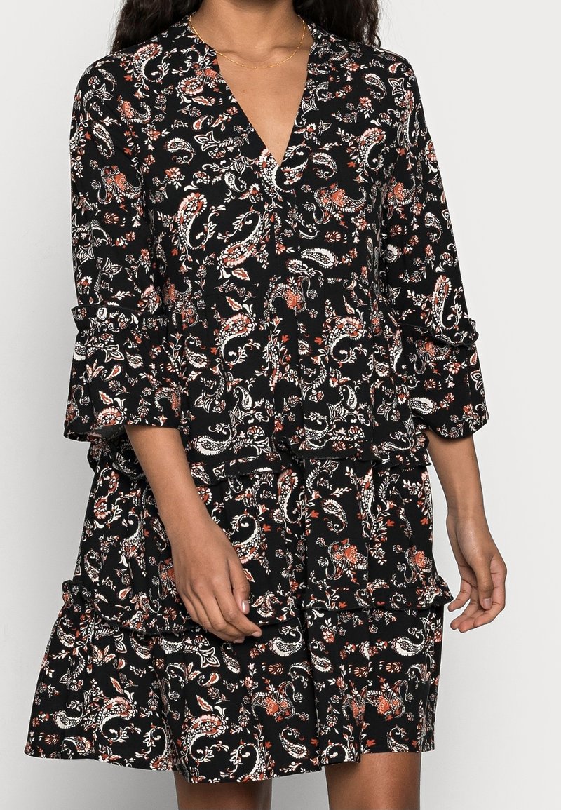 Femme portant une robe noire avec des motifs floraux cachemire blancs et orange, avec un col en V et des manches volantées trois quarts.