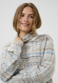 Młoda kobieta o jasnobrązowych włosach uśmiecha się, nosząc miękki, wielokolorowy sweter dziany z wysokim kołnierzem, delikatnie trzymając go blisko szyi.