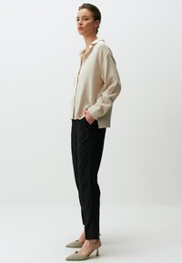 Beige strukturierte übergroße Bluse mit Kragen, schwarze schmal geschnittene Hose und hellfarbene spitze Absätze. Das Modell steht vor einem schlichten Hintergrund.