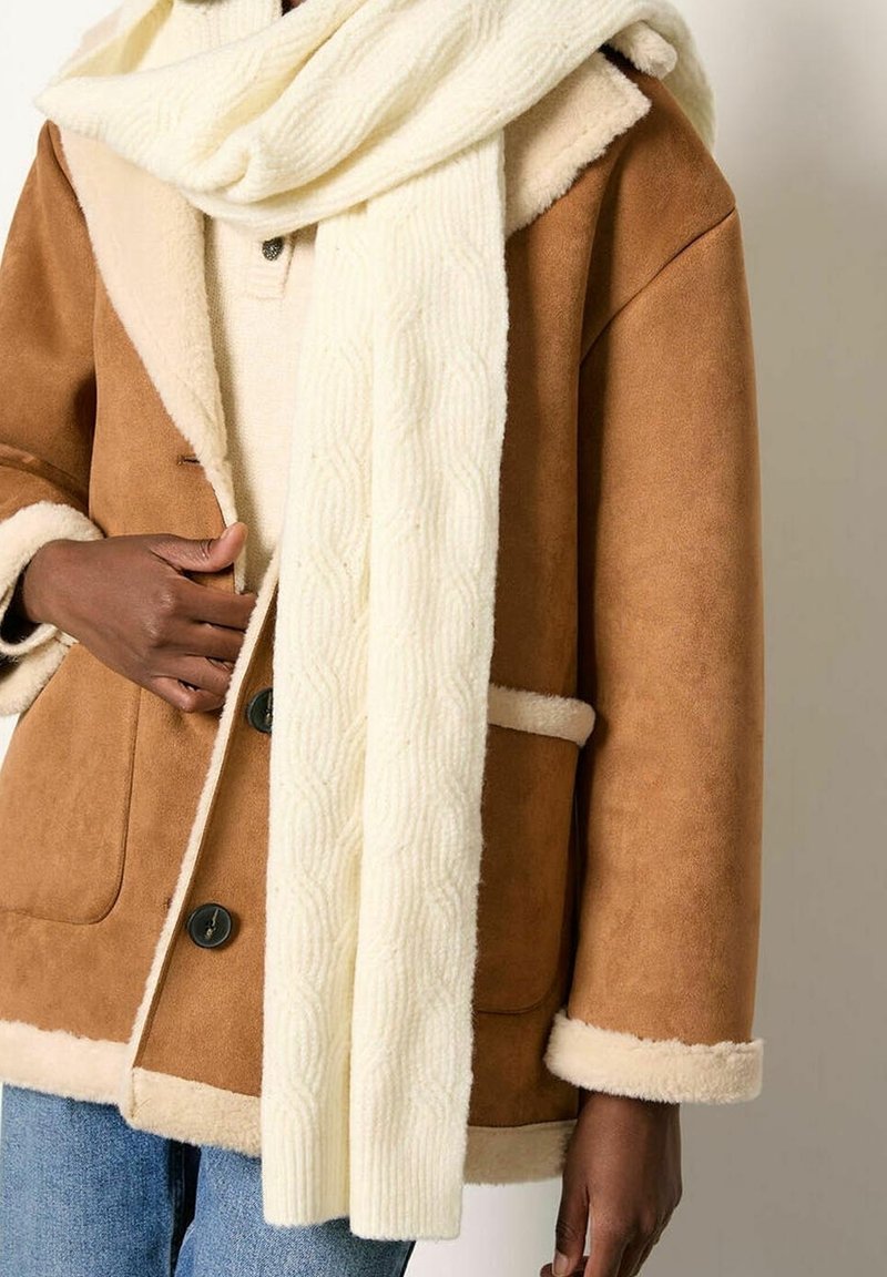 Personne portant un manteau beige doublé en peau lainée, une écharpe crème en tricot torsadé, un pull crème et un jean bleu, ajustant le manteau d'une main.