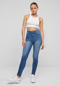 Pantalones skinny de cintura alta en azul, con una textura de denim suave, que presentan bolsillos frontales y un cierre de botón. Combinados con una camiseta cropped blanca y zapatillas.
