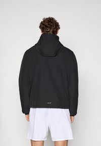 Veste à capuche noire avec une texture lisse, dotée d'un col haut et d'un détail de logo à l'arrière. Portée sur un short blanc.