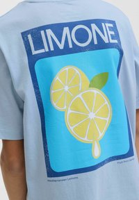 Šviesiai mėlyna medvilninė marškinėlė su grafiką, vaizduojančia dvi citrinos puseles mėlyname fone, viršuje esantis žodis "LIMONE" ir kelios lašelio sultys.