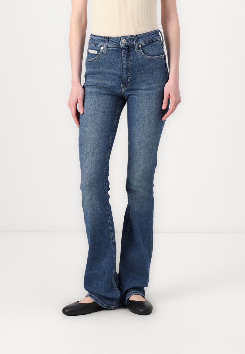 Jeans de mezclilla azul de corte acampanado con cintura alta, cinco bolsillos y un cierre de botón clásico. Textura suave con un ligero desvanecimiento.