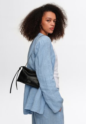 Kobieta z kręconymi włosami, ubrana w jasnoniebieski garnitur w prążki i biały top, nosząca małą czarną skórzaną torbę na ramię.