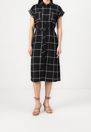 Zwarte midi overhemdjurk met wit windowpane ruitpatroon, korte mouwen, knoopsluiting voor, strikceintuur in de taille, gedragen met beige sandaalhakken met bandjes.