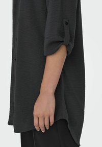 Donkergrijze shirt van een textuurstof, met opgerolde mouwen vastgemaakt met knopen, en een gebogen zoom. Het shirt is iets oversized.