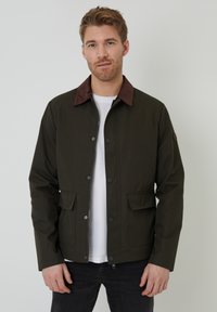 Veste vert foncé avec un col en velours côtelé marron, boutons-pression, deux poches avant et une coupe droite. Portée sur une chemise blanche.