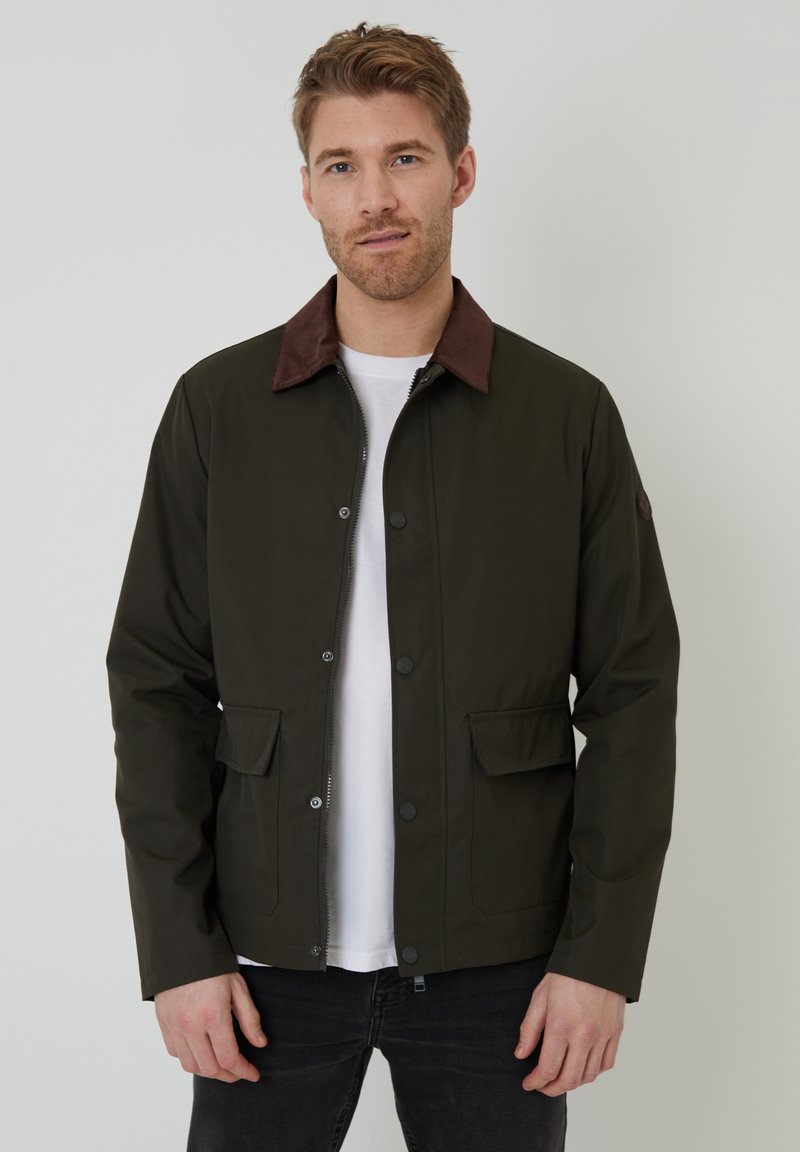 Veste vert foncé avec un col en velours côtelé marron, boutons-pression, deux poches avant et une coupe droite. Portée sur une chemise blanche.