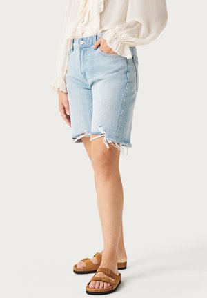 Mujer con pantalones cortos de mezclilla desgastada en azul claro, una blusa blanca de manga larga y sandalias marrones de doble correa, de pie con una mano en el bolsillo.