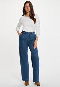 Top blanco de manga larga con detalle decorativo en el cuello, combinado con jeans de pierna ancha azules de cintura alta y botas marrones hasta el tobillo.