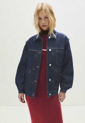 Blonde vrouw in een donkerblauwe denimjack over een rode jurk, staand tegen een eenvoudige lichte achtergrond.
