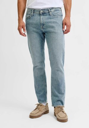 Un uomo che indossa jeans di denim blu chiaro, scarpe beige casual con lacci e calzini bianchi, in piedi davanti a uno sfondo bianco.