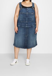 Džínová šaty s laclem v tmavě modré barvě, se stahovacími ramínky, přední kapsou a volným střihem. V kombinaci s bílými teniskami.
