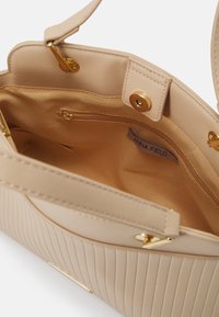 Beige käsilaukku, jossa on uritettu pinta, kultaiset yksityiskohdat ja kangaspintaan. Sisällä on vetoketjullinen tasku ja etiketti, jossa lukee "ANNA FIELD."