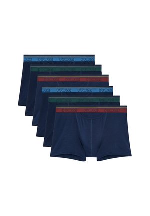 Fitte man staat blootsvoets en draagt donkerblauwe boxershorts tegen een effen lichte achtergrond.