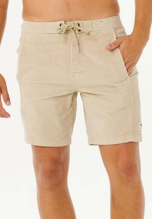 Beige shorts met trekkoord gedragen door een persoon met één hand in de zak en een zichtbare tatoeage op de bovenarm.