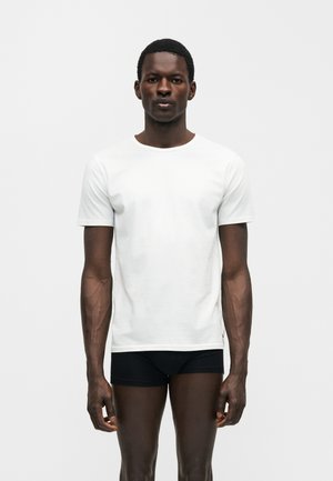 Mand, der står og vender fremad iført en ensfarvet hvid kortærmet T-shirt og sort undertøj mod en neutral baggrund.