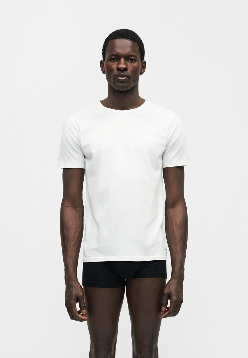 Homme debout, faisant face, portant un t-shirt blanc uni à manches courtes et un sous-vêtement noir sur un fond neutre.