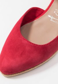 Chaussure slip-on en suède rouge avec un bout pointu, présentant un design découpé sur les côtés et une semelle en jute texturée.