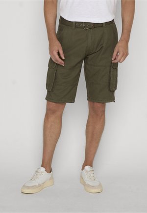 Mand iført olivengrønne cargoshorts, hvidt bælte og hvide sneakers, stående mod en ensartet lys baggrund.