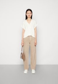 Weißes Kabelstrick-Cardigan mit kurzen Ärmeln und Knopfverschluss, kombiniert mit beigen weit geschnittenen Hosen und weißen Turnschuhen. Braune Handtasche inklusive.