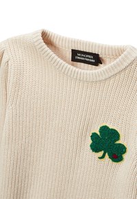 Pull en tricot beige avec un col en côtes. Présente un patch en feutre vert en forme de trèfle avec un détail en cœur rouge sur la poitrine. Étiquette de marque visible.