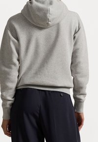 Sudadera gris con capucha, con un tejido texturizado y dobladillo acanalado, combinada con pantalones oscuros que tienen bolsillos laterales y detalles de botones.