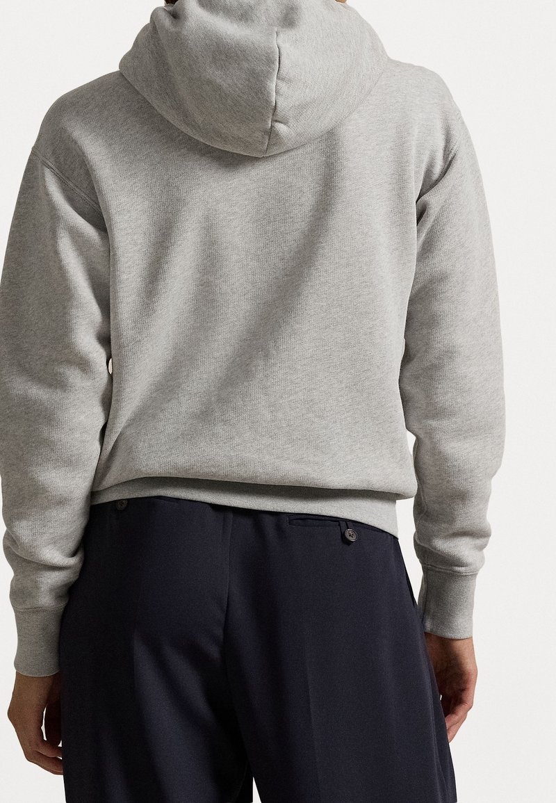 Sudadera gris con capucha, con un tejido texturizado y dobladillo acanalado, combinada con pantalones oscuros que tienen bolsillos laterales y detalles de botones.