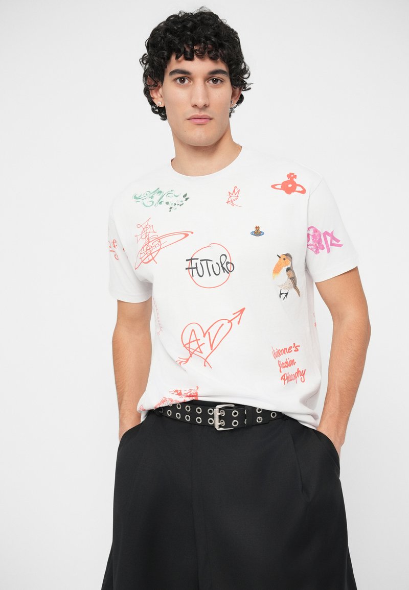 Jeune homme aux cheveux bouclés portant un t-shirt graphique blanc avec des dessins rouges, un oiseau et le mot "FUTURO", associé à un pantalon noir et une ceinture à œillets.