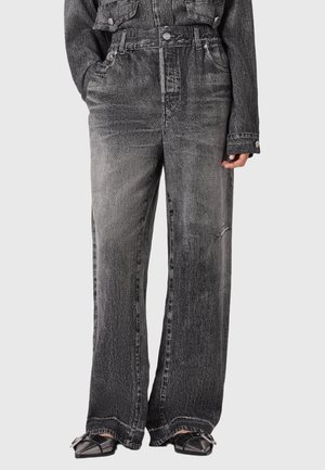 Kobieta nosząca czarne, dopasowane w talii, luźne jeansy z efektem prania kwasowego i przednimi kieszeniami, zestawione z czarnymi, szpiczastymi butami z srebrnymi klamrami.