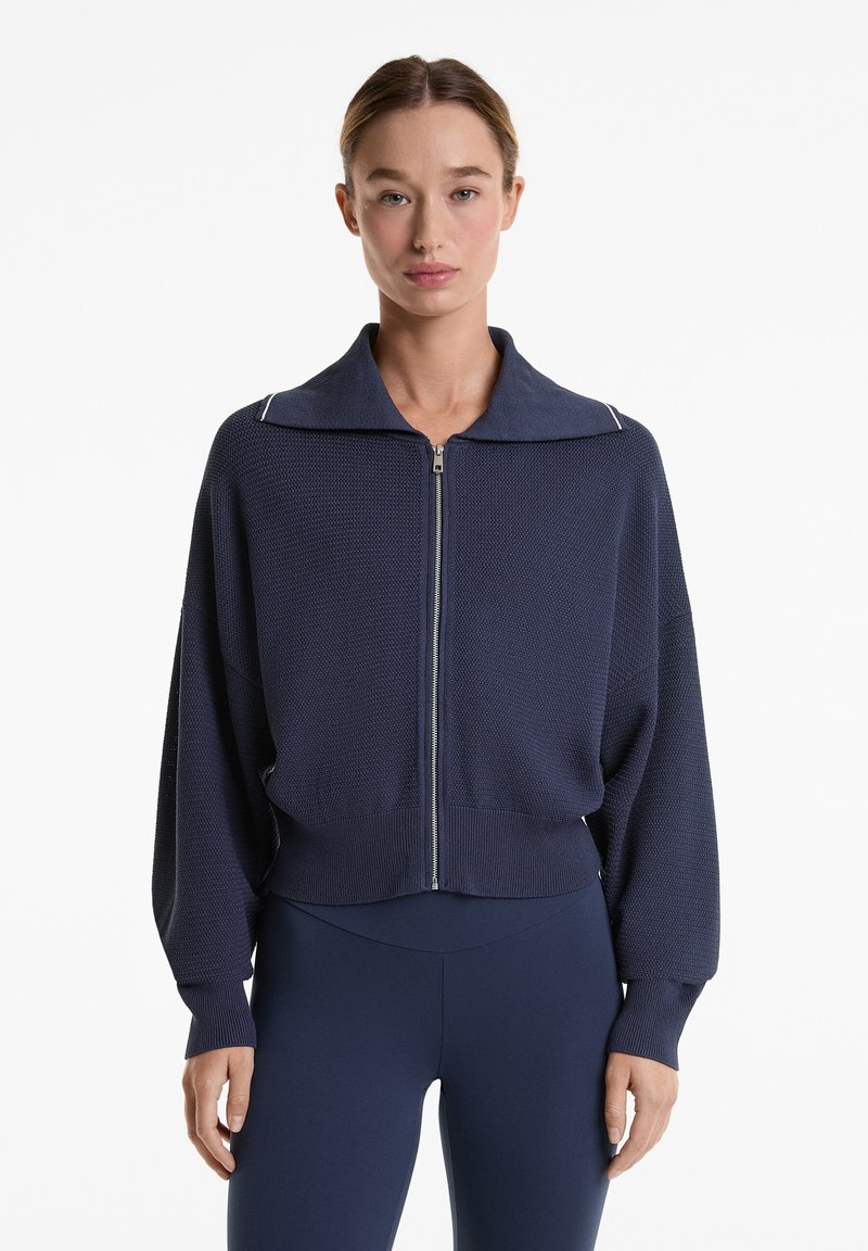 Navyblauer, verkürzter Reißverschluss-Pullover mit weitem Kragen, strukturiertem Stoff und weißen Akzentnähte. Lange, lockere Ärmel mit gerippten Bündchen.