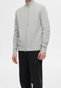 Ljusgrå zip-up tröja med hög krage, strukturerad stickad design, ribbade ärmslut och nederkant, bärs över mörka byxor.