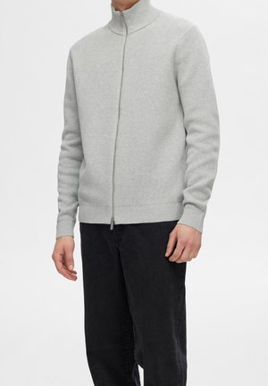 Lichtgrijze zip-up trui met een hoge kraag, textuur gebreid patroon, ribbelmanchetten en -zoom, gedragen over donkere broek.