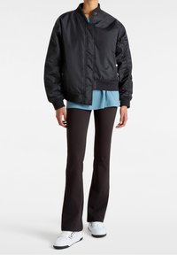 Blouson aviateur noir avec une texture lisse et brillante ; poignets et ourlet côtelés ; superposé sur un haut bleu clair ; associé à un pantalon évasé noir et des baskets blanches.