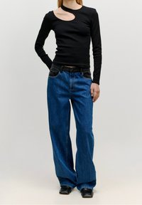Schwarzes Langarmoberteil mit einem runden Ausschnitt, kombiniert mit lockeren blauen Jeans aus Denim, die einen schwarzen Akzent am Bund und große Taschen haben.
