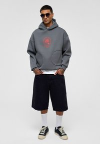 Grijze hoodie met een rode roosgrafiek, met grote voorzakken en een relaxte pasvorm. Draagt met zwarte shorts en sneakers.