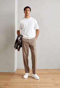 Hvit t-skjorte med korte ermer, lys brune skreddersydde bukser og hvite sneakers. Grå rutete flanelljakke over armen. Bakgrunn med tregulv.
