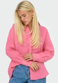 Camicetta rosa con bottoni, dalla vestibilità comoda, maniche lunghe e texture morbida; abbinata a jeans blu a vita alta.