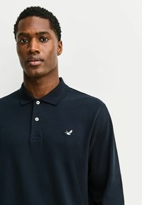 Polo azul marino con cuello, que presenta una tapeta de tres botones y un pequeño bordado de un pájaro en el pecho izquierdo. Tela de algodón, textura suave.