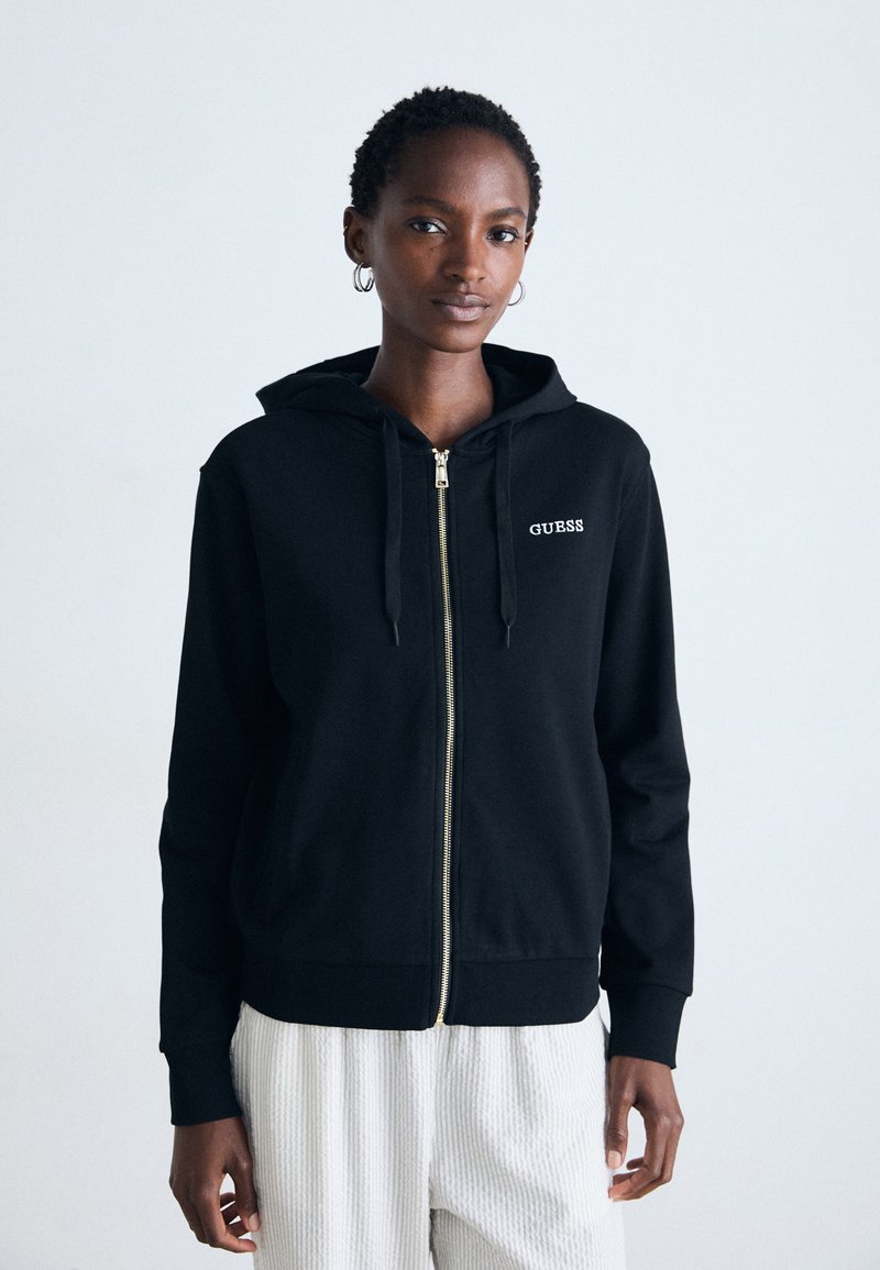 Vrouw in een zwarte hoodie met rits en het "GUESS" logo, met witte geribbelde broek, staand tegen een effen lichte achtergrond.
