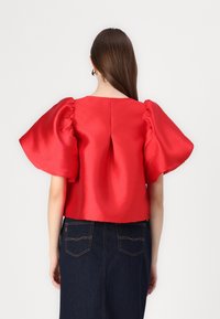 Blusa rossa in raso a maniche corte con maniche a palloncino oversize e scollo quadrato. Tessuto opaco con superficie liscia, indossata sopra una gonna in denim scuro.
