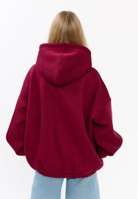 Felpa oversize color bordeaux con un grande cappuccio e vita elasticizzata, realizzata in materiale morbido. Presenta spalle scese e una vestibilità rilassata.