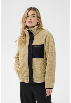 Ung kvinde iført beige fleecejakke med sort brystlomme og sorte bukser, stående med hænderne i jakkelommerne.