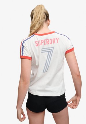 Kvinde med blond hestehale iført en hvid Superdry T-shirt med røde kanter og nummer 7, kombineret med sorte shorts, vendt væk fra kameraet.