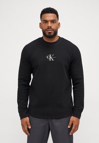 Zwarte lange mouw trui van een textuurstof met een Calvin Klein-logo op de borst. Geribbelde boorden en ontwerp met ronde hals.