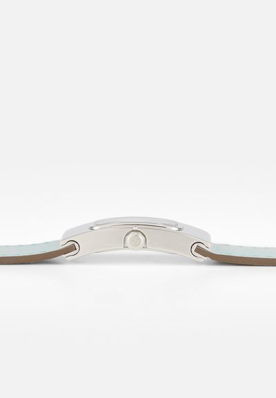 Silbernes rechteckiges Uhrgehäuse Seitenansicht mit Krone, befestigt an hellblauen Lederarmbändern auf weißem Hintergrund.