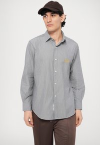 Srajca z dolgimi rokavi s sivimi in belimi črtami, iz lahkega materiala, z ukrivljenim spodnjim robom in subtilnim logotipom na prsih.