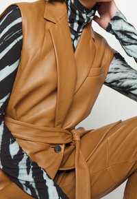 Person iført en tan lædervest og bukser med en sort-hvid mønstret turtleneck, poserer med hånden nær ansigtet.