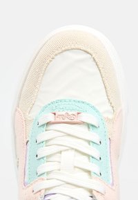 Gros plan du bout de la chaussure de sport avec des lacets blancs, des panneaux en tissu rose pastel, beige et vert menthe, et une petite étiquette rose sur les lacets.
